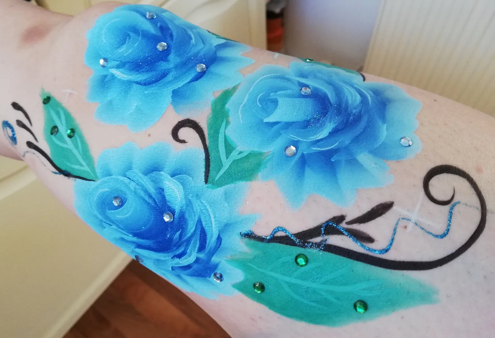Drei blaue Rosen mit grünen Blättern, Ranken und aufgeklebten Strasssteinen, auf den Unterarm einer Erwachsenen gemalt.
