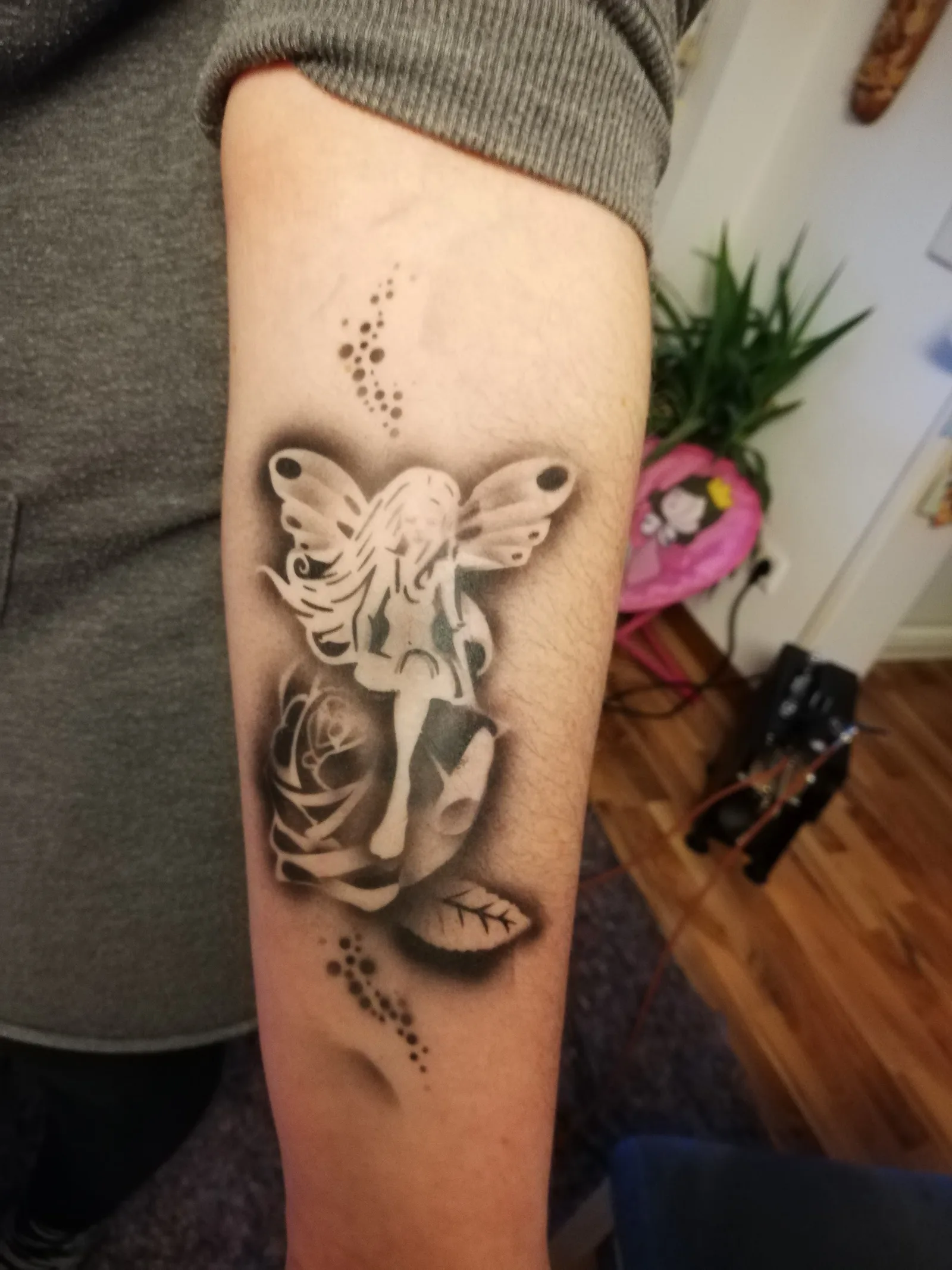 Airbrushtattoo auf einem Unterarm: eine Elfe mit Flügeln über einer Rose, dahinter der Arbeitsplatz mit der Airbrush-Pistole.