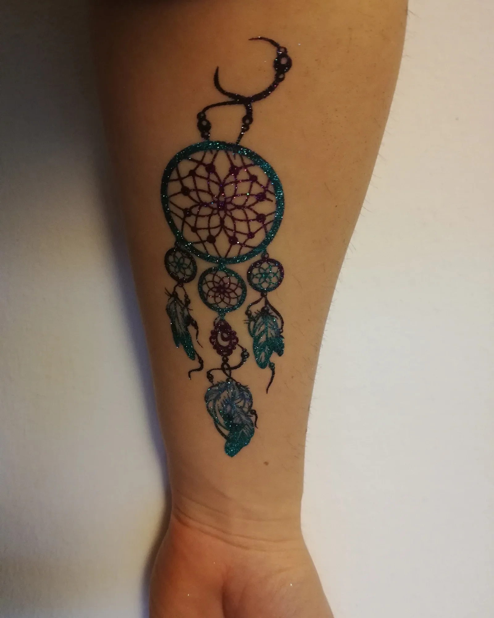 Glitzertattoo eines Traumfängers in Türkis und Violett mit drei Federn auf einem Unterarm.