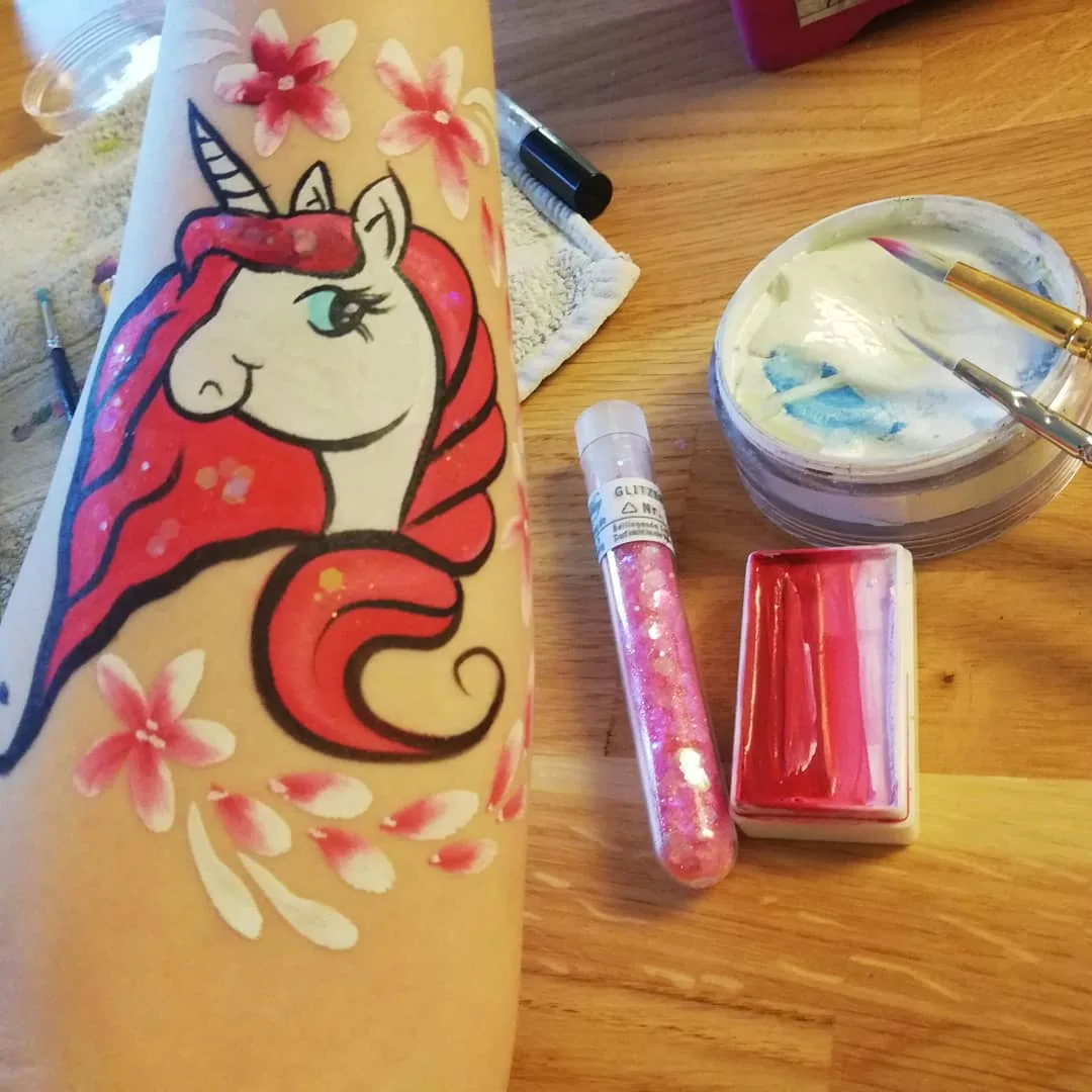 Ein rosa Einhorn mit Blüten, auf einen Arm gemalt, daneben auf dem Holztisch die Schminkfarben, zwei Pinsel und ein Röhrchen Glitzer.
