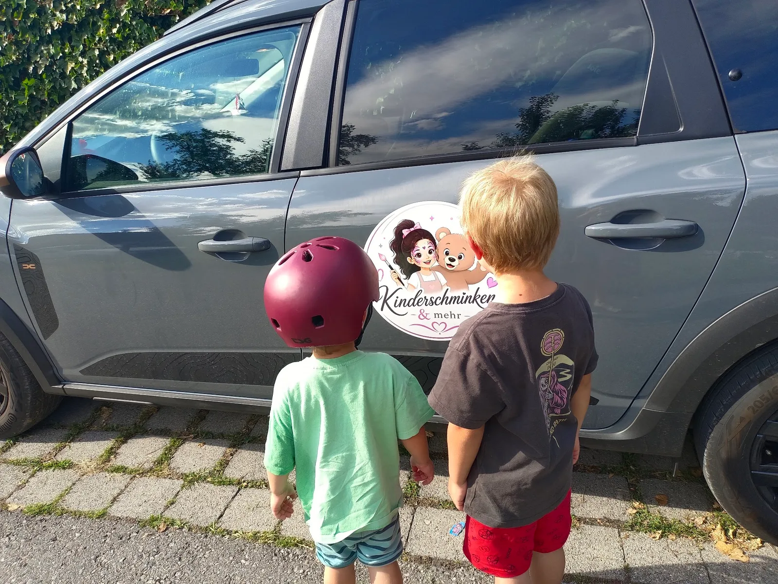 Ein grauer Kombi mit der runden Magnettafel „Kinderschminken & mehr“ an der Fahrertür, davor zwei Kinder von hinten.